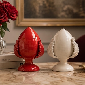 Può includere: Due sculture decorative in ceramica, una rossa e una bianca. Entrambe hanno una forma a goccia con dettagli a forma di foglie. Sono posizionate su una superficie di marmo, con un vaso di rose rosse sullo sfondo.