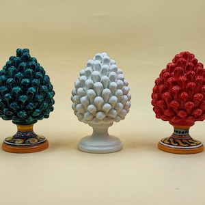 May include: Three ceramic pinecone sculptures in teal, white, and red. Each has a detailed, textured surface and sits on a decorative base with a painted design. The sculptures are displayed against a neutral background.