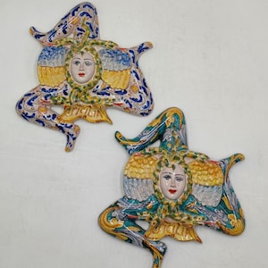 Può includere: Due figure in ceramica della Trinacria, un simbolo tradizionale siciliano. Ogni figura presenta un volto femminile con labbra rosse, circondato da dettagli gialli, blu e verdi. Le figure hanno tre gambe e sono decorate con motivi floreali.
