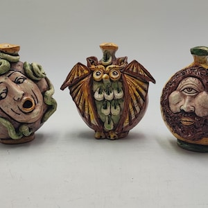 Op de afbeelding: Drie keramische decoratieve flessen met unieke gezichtsontwerpen. Eén toont Medusa met groene slangen, een andere een uil met vleugels en de laatste een bebaard gezicht. Elke fles heeft een ander kleurenschema.