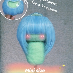 Puede incluir: Una cabeza de muñeca de tamaño mini con cabello azul claro y un cuerpo verde. El texto dice "Compartimento para un llavero" y "Tamaño mini Alrededor de la cabeza 12,7 cm - 14 cm". Tiene un compartimento para llavero.