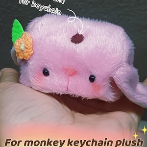 Könnte beinhalten: Ein rosa Plüsch-Affen-Schlüsselanhänger mit einem Fach für einen Schlüsselanhänger. Das Plüschtier hat eine Blumen- und Blattverzierung auf dem Kopf und ist 17 cm lang.