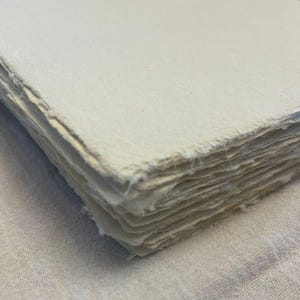 May include: A stack of off-white handmade paper sheets with rough, deckled edges. The paper has a slightly textured surface and is neatly stacked. The paper is likely used for art, calligraphy, or journaling.