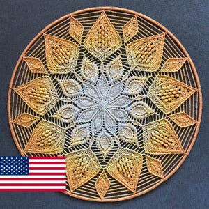 Może przedstawiać: Okrągła, ręcznie robiona serwetka z precyzyjnym, symetrycznym wzorem. Serwetka ma gradient kolorów, przechodzący od jasnokremowego środka do odcieni żółtego i pomarańczowego na krawędziach. Złożony wzór zawiera kształty rombów i płatków.