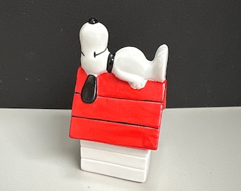 May include: A white ceramic figurine of Snoopy, the beagle from the Peanuts comic strip, lying on top of his red doghouse.