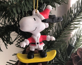 Puede incluir: Un adorno navideño de Snoopy. Snoopy lleva un gorro de Papá Noel y una chaqueta roja con ribete blanco, montando en una tabla de snowboard amarilla. El adorno cuelga de un hilo dorado, sobre un fondo de árbol de Navidad.
