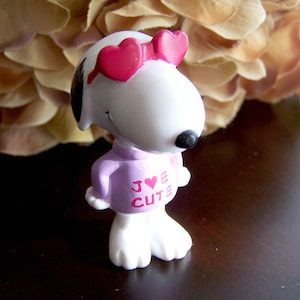 May include: A white Snoopy figurine wearing pink heart-shaped sunglasses and a pink shirt with the words "I ♥ CUTE" printed on it.