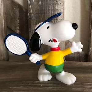 Può includere: Una statuetta bianca di Snoopy con un orecchio nero, che indossa una maglietta gialla, pantaloncini verdi, un collare rosso e una visiera da tennis blu. Il personaggio tiene una racchetta da tennis blu e saluta con l'altra mano.