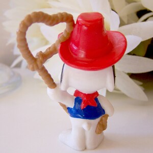 Pu&ograve; includere: Una statuina di Snoopy vestito da cowboy, vista da dietro. La figura indossa un cappello da cowboy rosso, un gilet blu e un fazzoletto rosso. Tiene un lazo marrone nella mano destra. La statuina &egrave; bianca con orecchie nere.