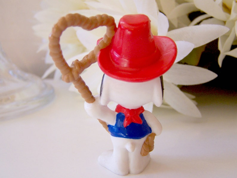 Pu&ograve; includere: Una statuina di Snoopy vestito da cowboy, vista da dietro. La figura indossa un cappello da cowboy rosso, un gilet blu e un fazzoletto rosso. Tiene un lazo marrone nella mano destra. La statuina &egrave; bianca con orecchie nere.