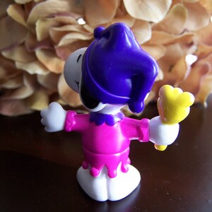 May include: A purple and pink plastic toy figure of a jester with a yellow wand. The figure is wearing a purple hat with a jester's bell.