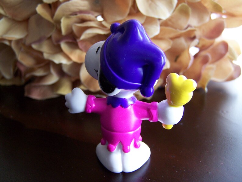 May include: A purple and pink plastic toy figure of a jester with a yellow wand. The figure is wearing a purple hat with a jester's bell.