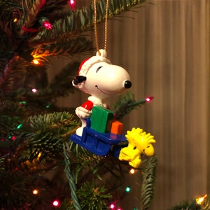 Puede incluir: Un adorno navideño blanco y rojo con Snoopy vestido de Papá Noel tirando de un trineo azul con Woodstock. El adorno está colgado de un árbol de Navidad verde con luces de colores.