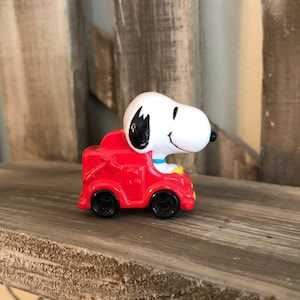 Può includere: Un camioncino dei pompieri rosso giocattolo con una figura bianca di Snoopy al volante. Snoopy indossa un collare blu e ha gli occhi neri.