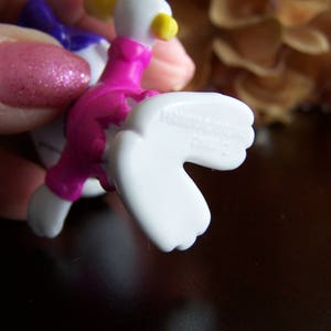 May include: A pink and white plastic toy duck with a yellow beak and a purple bow. The duck has a white wing that is outstretched.
