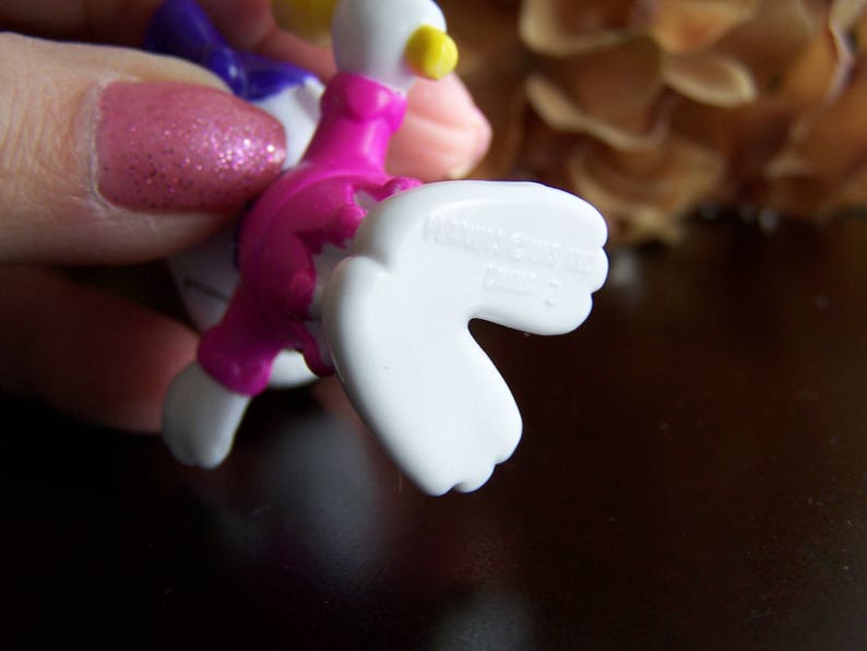 May include: A pink and white plastic toy duck with a yellow beak and a purple bow. The duck has a white wing that is outstretched.