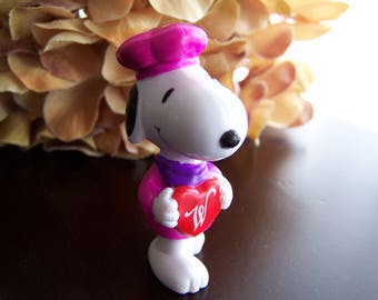 Puede incluir: Una figurita de plástico blanca de un personaje de dibujos animados que lleva un gorro de cocinero rosa y una bufanda morada. El personaje sostiene un corazón rojo con una letra 'W' blanca.