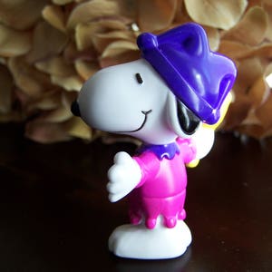 May include: A white and purple cartoon character with a purple hat and a pink dress. The character is holding a yellow object in its right hand.
