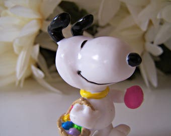 Puede incluir: Una figurita de plástico blanca de un perro de dibujos animados con orejas negras y nariz negra. El perro sostiene una cesta con huevos de Pascua coloridos.