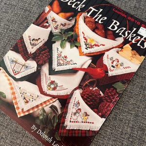 Puede incluir: Una portada de libro titulada "Deck The Baskets" de Deborah Lambein. La portada presenta diseños de punto de cruz de cestas temáticas navideñas, incluyendo a Papá Noel, muñecos de nieve y arreglos florales festivos. Los diseños se presentan sobre tela a cuadros y blanca.