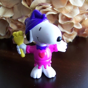 May include: A white plastic toy figure of Snoopy dressed as a jester. He is wearing a purple hat with a yellow and orange jester's wand in his right hand. He is wearing a pink and purple costume with a heart on the chest.