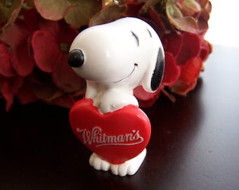 Puede incluir: Una figurita de cerámica blanca de Snoopy sosteniendo un corazón rojo con el texto "Whitman's" en letras blancas. La figurita está sentada sobre una superficie marrón oscura.