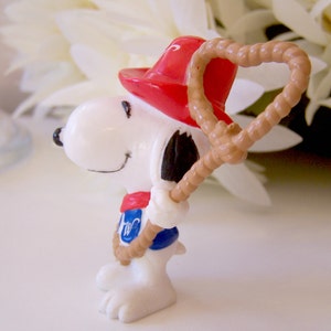 Pu&ograve; includere: Una piccola statuetta bianca di un cane dei cartoni animati che indossa un cappello rosso, un fazzoletto rosso e un gilet blu con una lettera W bianca. Il cane tiene in mano un lazo di corda color marrone chiaro. Orecchie, naso e bocca neri.