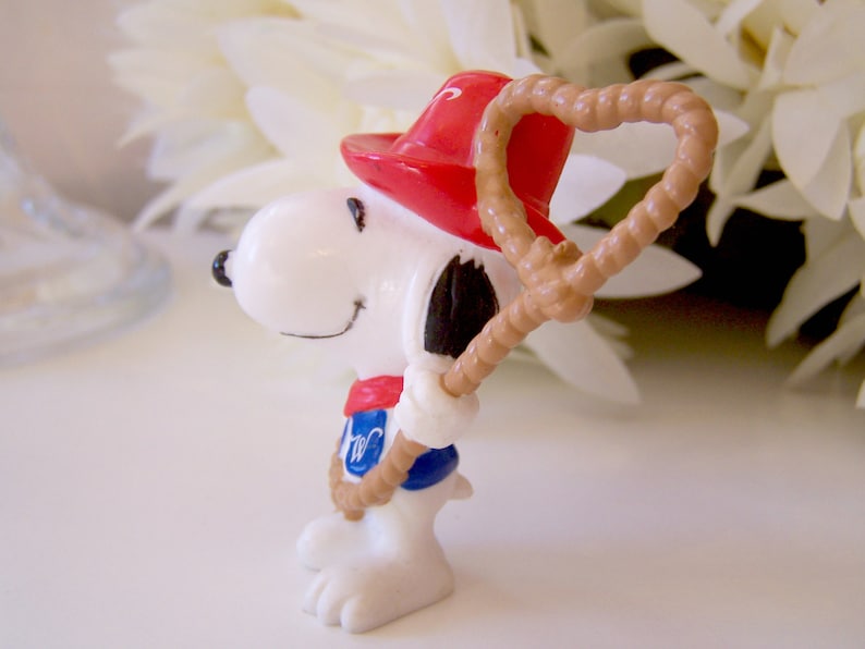 Pu&ograve; includere: Una piccola statuetta bianca di un cane dei cartoni animati che indossa un cappello rosso, un fazzoletto rosso e un gilet blu con una lettera W bianca. Il cane tiene in mano un lazo di corda color marrone chiaro. Orecchie, naso e bocca neri.