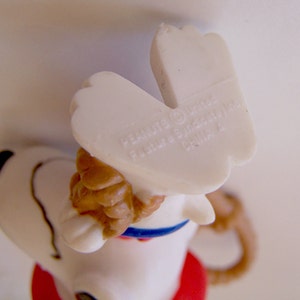 Pu&ograve; includere: Una statuetta in plastica bianca di un cane con orecchie marroni, un collare blu e una base rossa. La pianta dei piedi presenta il testo stampato PEANUTS (c) United Feature Syndicate, Inc. CHINA 4, visibile dall'alto.