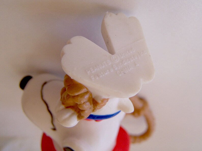 Pu&ograve; includere: Una statuetta in plastica bianca di un cane con orecchie marroni, un collare blu e una base rossa. La pianta dei piedi presenta il testo stampato PEANUTS (c) United Feature Syndicate, Inc. CHINA 4, visibile dall'alto.