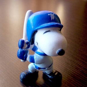 May include: A small Snoopy figurine dressed in a white and blue baseball uniform, wearing a blue batting helmet with a white letter W on the front. The character is holding a white baseball bat, has black ears, and black shoes.