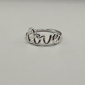 May include: A silver ring with the word "love" in a cursive script. The ring is made of a shiny, reflective metal and sits on a plain white surface. The word "love" is the focal point of the ring's design.