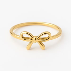 May include: A gold-colored ring featuring a bow design. The ring has a thin band and a three-dimensional bow in the center. The bow has a detailed, open design. The ring is set against a white background.