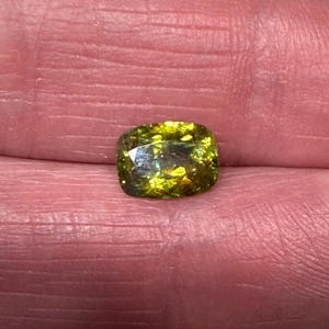 May include: A cushion-cut gemstone with a mix of green and yellow hues. The gem has a faceted surface that reflects light, creating a sparkling effect. The stone is held in a hand, showcasing its size and color.