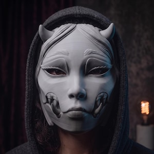 Puede incluir: Una máscara de color gris claro con textura lisa que presenta rasgos faciales estilizados, dos pequeños cuernos en la frente y colmillos prominentes que sobresalen de las mejillas. La máscara se lleva sobre el rostro, enmarcada por una sudadera gris oscuro.