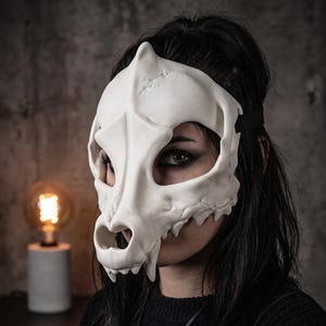 Peut inclure: Un masque en forme de crâne d'animal blanc moulé, doté d'un museau pointu, de grandes ouvertures pour les yeux et de dents visibles le long de la mâchoire. Le masque est maintenu par une sangle élastique noire qui entoure la tête.