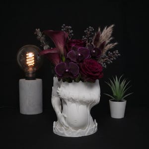 Könnte beinhalten: Eine weiße Vase mit einem skulpturalen, fließenden, handartigen Design, gefüllt mit einem Strauß aus dunkelvioletten Calla-Lilien, tiefroten Rosen, dunklen Orchideen und getrockneten Gräsern. Links eine Glühbirne auf Marmorsockel, rechts eine kleine Sukkulente.