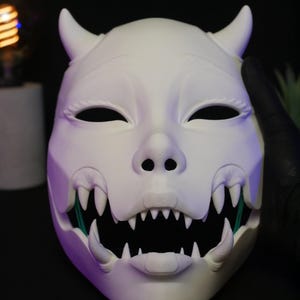 May include: A matte white mask featuring two curved horns at the top, almond-shaped eye openings, and a wide, open mouth displaying sharp, pointed teeth. The design includes a snout-like nose structure and visible fangs on the upper and lower jaw.