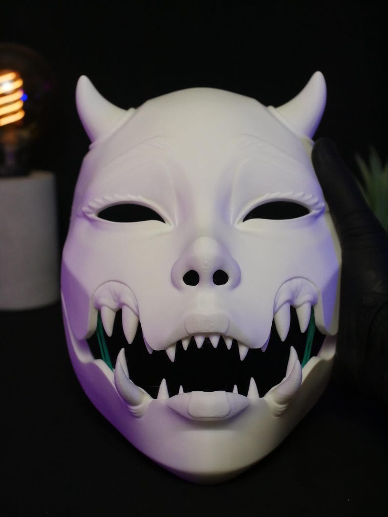 May include: A matte white mask featuring two curved horns at the top, almond-shaped eye openings, and a wide, open mouth displaying sharp, pointed teeth. The design includes a snout-like nose structure and visible fangs on the upper and lower jaw.