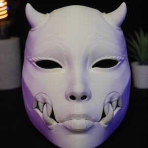 May include: A matte white mask featuring a stylized human face with two prominent horns on the forehead, almond-shaped eye openings, and a mouth area displaying sharp, protruding fangs on both sides. The mask has a smooth, solid surface.