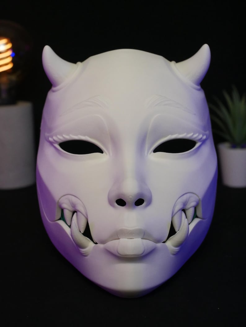 May include: A matte white mask featuring a stylized human face with two prominent horns on the forehead, almond-shaped eye openings, and a mouth area displaying sharp, protruding fangs on both sides. The mask has a smooth, solid surface.