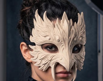 May include: A beige, sculpted mask featuring intricate, layered feather-like textures and two prominent downward-pointing fangs. The mask covers the upper face and is secured with a black elastic strap around the head.