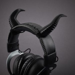 May include: Black over-ear headphones with a leather headband and two curved, textured horns attached. The horns and headband are stitched with white thread. The headphones are set against a grey background.