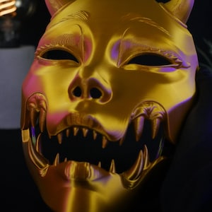 May include: A metallic gold-colored mask featuring two prominent horns at the top, almond-shaped eye openings, and a wide, open mouth displaying a set of sharp, pointed teeth. The surface has a smooth, reflective finish with subtle contours.
