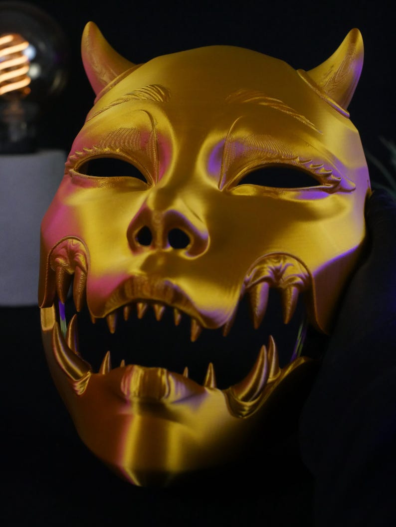 May include: A metallic gold-colored mask featuring two prominent horns at the top, almond-shaped eye openings, and a wide, open mouth displaying a set of sharp, pointed teeth. The surface has a smooth, reflective finish with subtle contours.