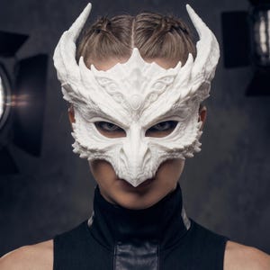 Op de afbeelding: Een wit, getextureerd masker geïnspireerd op een draak, met ingewikkelde schubpatronen, een puntige snavelachtige neus en twee prominente, gebogen hoorns die naar boven uitsteken. Het masker bedekt het bovenste deel van het gezicht en laat de ogen vrij.