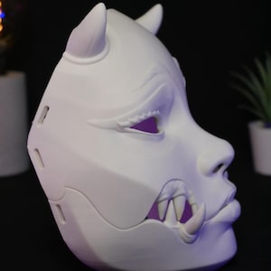May include: A matte white, three-dimensional mask featuring a stylized oni-inspired design. The mask displays prominent horns, sharp fangs protruding from the mouth area, and defined facial features including almond-shaped eyes and a nose.