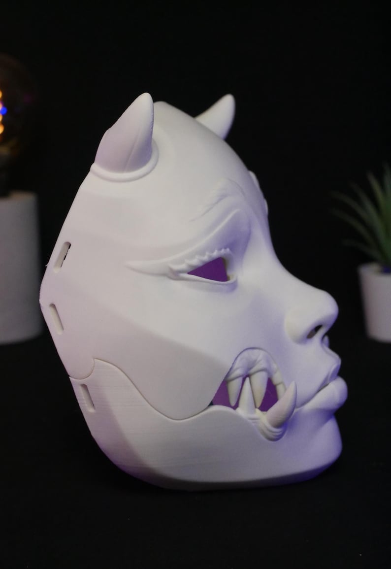 May include: A matte white, three-dimensional mask featuring a stylized oni-inspired design. The mask displays prominent horns, sharp fangs protruding from the mouth area, and defined facial features including almond-shaped eyes and a nose.