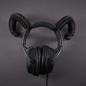 May include: Black headphones with a distinctive design, featuring curved, horn-like accents on the headband. The earcups are square and padded, connected by a silver wire. The headphones are set against a dark grey background.