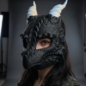 Peut inclure: Un masque de dragon noir avec une surface texturée rappelant des écailles, une crête centrale proéminente et deux cornes incurvées bleu clair sur le dessus. Le masque couvre le haut du visage, laissant les yeux visibles, et s'attache par une sangle.